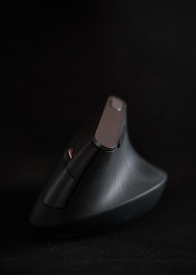 A black ergonomic computer mouse with a vertical design is set against a dark background. The mouse features a sleek, modern shape with multiple buttons visible. It appears angled slightly away, with a matte finish and a smooth texture.
