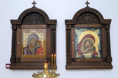 Two ornate iconographic paintings are housed in wooden frames with intricate craftsmanship and crosses at the top. Below the icons, several lit candles are placed on a decorative stand.