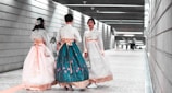 Three individuals wearing traditional garments walk through an underground corridor with modern architectural design, featuring linear lighting and tiled flooring. The garments are richly decorated and flow elegantly as they move. One person appears to be moving toward the viewer, while the other two walk away.