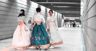 Three individuals wearing traditional garments walk through an underground corridor with modern architectural design, featuring linear lighting and tiled flooring. The garments are richly decorated and flow elegantly as they move. One person appears to be moving toward the viewer, while the other two walk away.