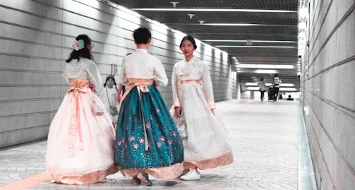 Three individuals wearing traditional garments walk through an underground corridor with modern architectural design, featuring linear lighting and tiled flooring. The garments are richly decorated and flow elegantly as they move. One person appears to be moving toward the viewer, while the other two walk away.