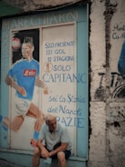 A mural painted on a textured wall depicts a soccer player in a blue jersey with statistics and accolades written in Italian. An older man with white hair sits pensively in front of the mural, holding a face mask. The scene has an urban and nostalgic feel.
