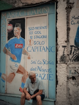 A mural painted on a textured wall depicts a soccer player in a blue jersey with statistics and accolades written in Italian. An older man with white hair sits pensively in front of the mural, holding a face mask. The scene has an urban and nostalgic feel.