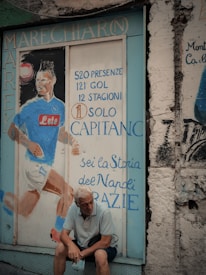 A mural painted on a textured wall depicts a soccer player in a blue jersey with statistics and accolades written in Italian. An older man with white hair sits pensively in front of the mural, holding a face mask. The scene has an urban and nostalgic feel.