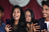 A group of young people are smiling and holding smartphones, appearing to be enjoying a joyful moment. They are captured in close proximity, suggesting a lively social setting. Their expressions reflect happiness and laughter.
