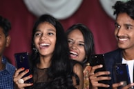 A group of young people are smiling and holding smartphones, appearing to be enjoying a joyful moment. They are captured in close proximity, suggesting a lively social setting. Their expressions reflect happiness and laughter.