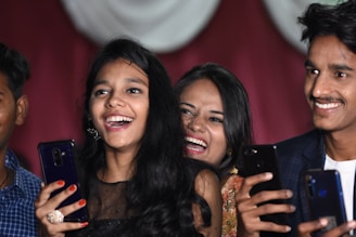 A group of young people are smiling and holding smartphones, appearing to be enjoying a joyful moment. They are captured in close proximity, suggesting a lively social setting. Their expressions reflect happiness and laughter.