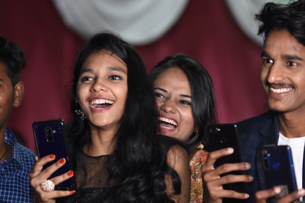 A group of young people are smiling and holding smartphones, appearing to be enjoying a joyful moment. They are captured in close proximity, suggesting a lively social setting. Their expressions reflect happiness and laughter.