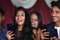 A group of young people are smiling and holding smartphones, appearing to be enjoying a joyful moment. They are captured in close proximity, suggesting a lively social setting. Their expressions reflect happiness and laughter.