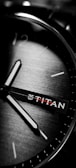 A close-up of a wristwatch with visible hour and minute hands on a brushed metallic dial. The brand name is visible, and the watch face features minimalistic hour markers.