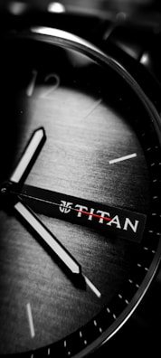 A close-up of a wristwatch with visible hour and minute hands on a brushed metallic dial. The brand name is visible, and the watch face features minimalistic hour markers.