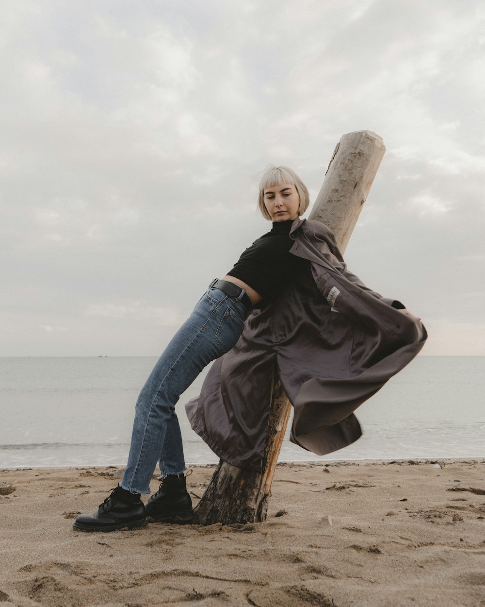 woman in black jacket and blue denim jeans sitting on brown wooden post near sea during