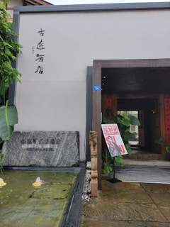 The entrance to a building has a modern architectural design, complemented by traditional elements such as a stone slab with engraved text. A small water feature with fountains provides a tranquil atmosphere. The entrance is adorned with some plants and a colorful sign board displaying Asian characters and vibrant graphics.