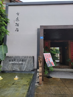 The entrance to a building has a modern architectural design, complemented by traditional elements such as a stone slab with engraved text. A small water feature with fountains provides a tranquil atmosphere. The entrance is adorned with some plants and a colorful sign board displaying Asian characters and vibrant graphics.