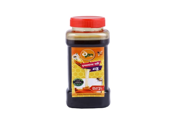 A clear plastic bottle with a red cap contains a dark liquid, likely honey, judging by the label design. The label features imagery of honeycombs and bees, indicating natural honey. The text on the label is in Bengali and includes the weight '500 gm.'