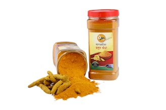 Two containers of turmeric powder are visible, one standing upright with a red lid and a label featuring an image of turmeric roots. The other container is lying on its side, spilling out the bright yellow spice next to a few whole turmeric roots.