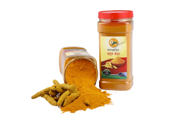 Two containers of turmeric powder are visible, one standing upright with a red lid and a label featuring an image of turmeric roots. The other container is lying on its side, spilling out the bright yellow spice next to a few whole turmeric roots.