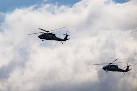 Two helicopters are flying in close formation against a backdrop of partly cloudy skies. The helicopters appear dark against the bright clouds.