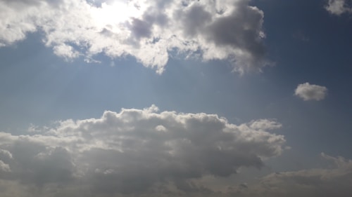 Bright clouds float in a vast blue sky, diffusing the sunlight and casting a gentle glow. Various shapes and densities of clouds provide a dynamic and serene scene.