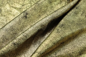 A close-up of a textured fabric with a golden, cracked pattern. The surface appears richly detailed, with varying shades of gold and black creating an intricate design. The fabric is layered and draped, showing folds and creases.