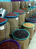 Multiple burlap sacks filled with various types of beans and grains are displayed. Each sack has a colored bowl on top with a scoop inside, and price tags are visible. The sacks are arranged in neat rows, showcasing a variety of colors and textures.