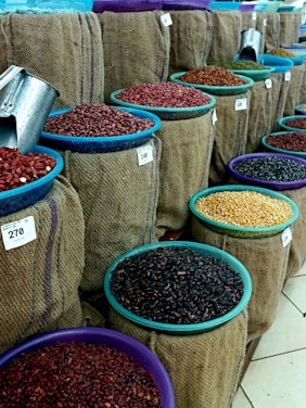 Multiple burlap sacks filled with various types of beans and grains are displayed. Each sack has a colored bowl on top with a scoop inside, and price tags are visible. The sacks are arranged in neat rows, showcasing a variety of colors and textures.