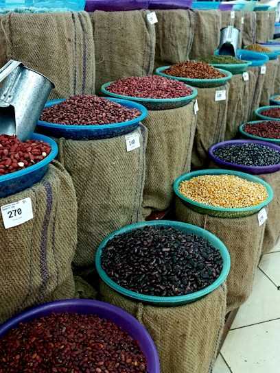 Multiple burlap sacks filled with various types of beans and grains are displayed. Each sack has a colored bowl on top with a scoop inside, and price tags are visible. The sacks are arranged in neat rows, showcasing a variety of colors and textures.