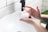 Hands applying a white cream from a jar labeled 'Ultra Hydrating Moisturiser' in a bathroom setting with a white sink and tiled walls.