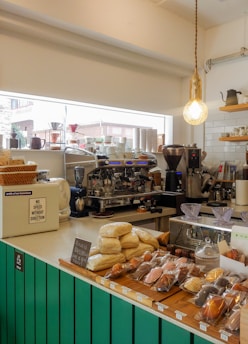 A cozy coffee shop counter featuring a large espresso machine surrounded by various coffee-making tools and accessories. Freshly baked goods are displayed at the front, including packaged pastries, buns, and bread. A warm light bulb hangs from the ceiling, casting a comforting glow. The counter is painted teal, and shelves in the background hold cups and other kitchenware.