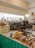 A cozy coffee shop counter featuring a large espresso machine surrounded by various coffee-making tools and accessories. Freshly baked goods are displayed at the front, including packaged pastries, buns, and bread. A warm light bulb hangs from the ceiling, casting a comforting glow. The counter is painted teal, and shelves in the background hold cups and other kitchenware.