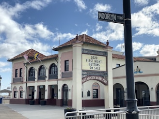 A vibrant building with a Mediterranean-style design, featuring arched windows and a colorful tile roof, located at the Ocean City Music Pier. Flags are displayed outside the building, and a sign advertises '2020 First Night Buttons On Sale.' The scene is well-lit with blue skies and scattered clouds above, and there is a street sign labeled 'MOORLYN TERRACE.'