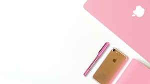A pink-themed flat lay with a pink laptop cover, a pink pen, a gold smartphone, and a pink notebook. The apple logo is visible on the laptop cover, indicating it is an Apple product.