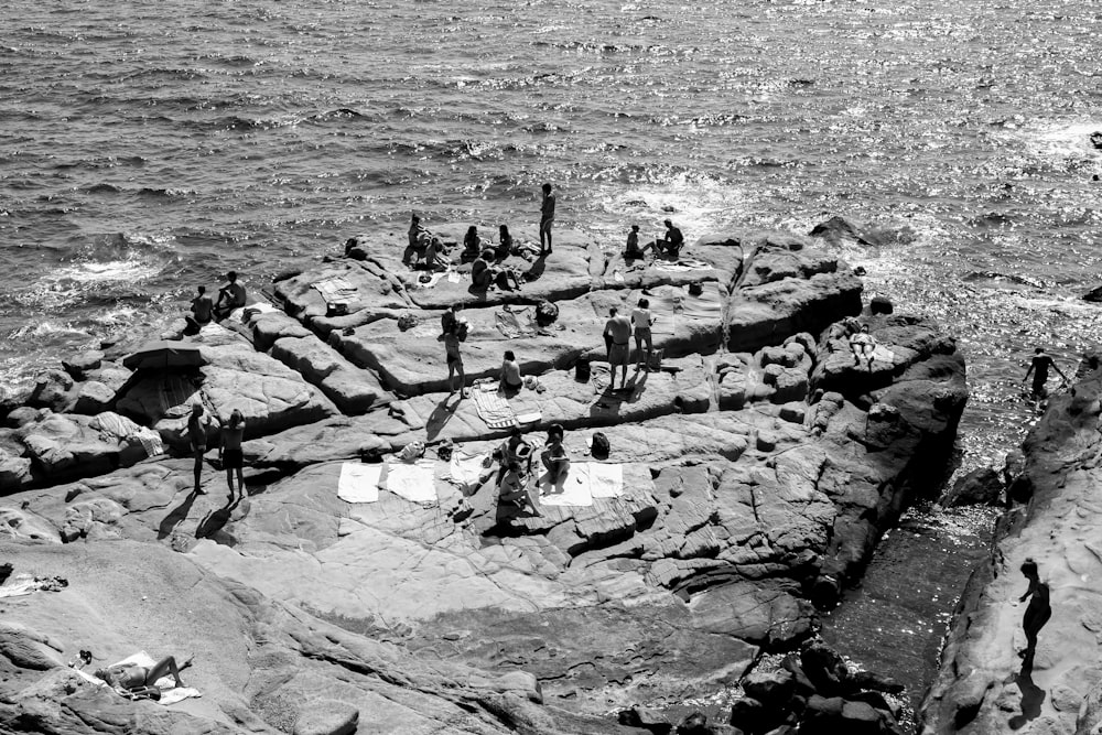 Foto en escala de grises de personas en una formación rocosa cerca del cuerpo de agua