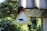 A triangular sign with a silhouette of an adult cat followed by three kittens is mounted under the edge of a building roof. The surroundings include lush green foliage and a blurred background of more vegetation.