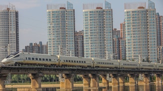 A high-speed train travels on a bridge across a river, set against a backdrop of modern high-rise buildings. The train is sleek and aerodynamic, moving along a well-constructed railway. The skyscrapers are predominantly glass and steel, with multiple windows reflecting daylight.