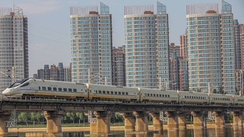 A high-speed train travels on a bridge across a river, set against a backdrop of modern high-rise buildings. The train is sleek and aerodynamic, moving along a well-constructed railway. The skyscrapers are predominantly glass and steel, with multiple windows reflecting daylight.