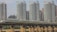 A high-speed train travels on a bridge across a river, set against a backdrop of modern high-rise buildings. The train is sleek and aerodynamic, moving along a well-constructed railway. The skyscrapers are predominantly glass and steel, with multiple windows reflecting daylight.