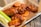 A tray filled with several pieces of cooked chicken wings covered in a glossy barbecue sauce, accompanied by fresh celery sticks. The tray is lined with brown parchment paper and rests on a wooden surface.