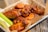 A tray filled with several pieces of cooked chicken wings covered in a glossy barbecue sauce, accompanied by fresh celery sticks. The tray is lined with brown parchment paper and rests on a wooden surface.