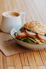 A close-up of a sandwich stacked with fresh vegetables and mozzarella cheese, topped with a crusty bun, placed on a light-colored ceramic plate. A cappuccino with frothy milk art is in a white mug beside the sandwich. The setup is on a wooden table with a branded napkin underneath.