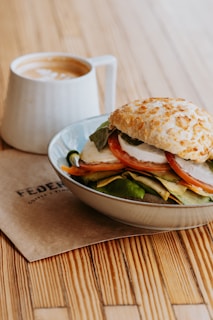 A close-up of a sandwich stacked with fresh vegetables and mozzarella cheese, topped with a crusty bun, placed on a light-colored ceramic plate. A cappuccino with frothy milk art is in a white mug beside the sandwich. The setup is on a wooden table with a branded napkin underneath.