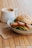 A close-up of a sandwich stacked with fresh vegetables and mozzarella cheese, topped with a crusty bun, placed on a light-colored ceramic plate. A cappuccino with frothy milk art is in a white mug beside the sandwich. The setup is on a wooden table with a branded napkin underneath.