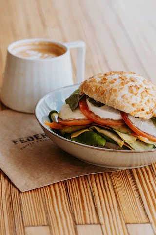 Close-up of a frothy cappuccino beside a freshly baked sandwich on cream-colored table.
