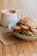 A close-up of a sandwich stacked with fresh vegetables and mozzarella cheese, topped with a crusty bun, placed on a light-colored ceramic plate. A cappuccino with frothy milk art is in a white mug beside the sandwich. The setup is on a wooden table with a branded napkin underneath.
