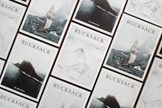 A collection of Rucksack magazine covers is arranged in a grid-like pattern. Each cover features a different dramatic landscape, including a sailing ship on a stormy sea, a rugged island, and snowy mountains. The covers are aesthetically coordinated, with a minimalist and adventurous theme.