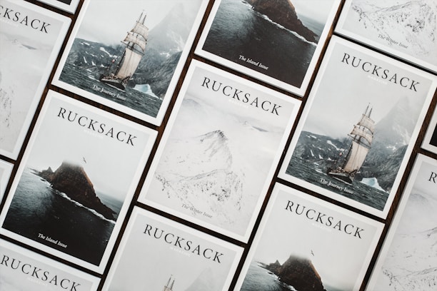 A collection of Rucksack magazine covers is arranged in a grid-like pattern. Each cover features a different dramatic landscape, including a sailing ship on a stormy sea, a rugged island, and snowy mountains. The covers are aesthetically coordinated, with a minimalist and adventurous theme.