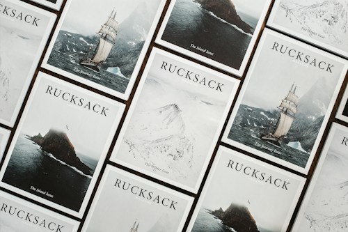 A collection of Rucksack magazine covers is arranged in a grid-like pattern. Each cover features a different dramatic landscape, including a sailing ship on a stormy sea, a rugged island, and snowy mountains. The covers are aesthetically coordinated, with a minimalist and adventurous theme.