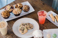 A variety of pastries and desserts are arranged on plates atop a rustic wooden table. There are muffins with different toppings and flavors, cookies, a cannoli, and small macarons. Two beverages in plastic cups, one iced coffee and another with a pink hue and fruit slices, are also visible.