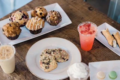 A variety of pastries and desserts are arranged on plates atop a rustic wooden table. There are muffins with different toppings and flavors, cookies, a cannoli, and small macarons. Two beverages in plastic cups, one iced coffee and another with a pink hue and fruit slices, are also visible.