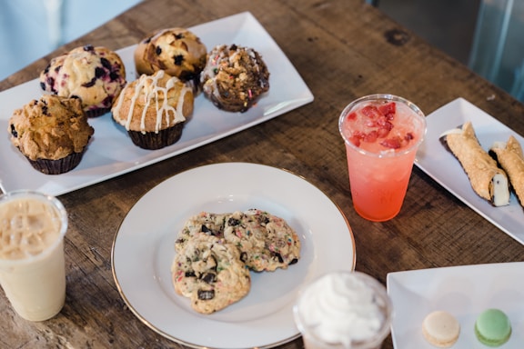 A variety of pastries and desserts are arranged on plates atop a rustic wooden table. There are muffins with different toppings and flavors, cookies, a cannoli, and small macarons. Two beverages in plastic cups, one iced coffee and another with a pink hue and fruit slices, are also visible.